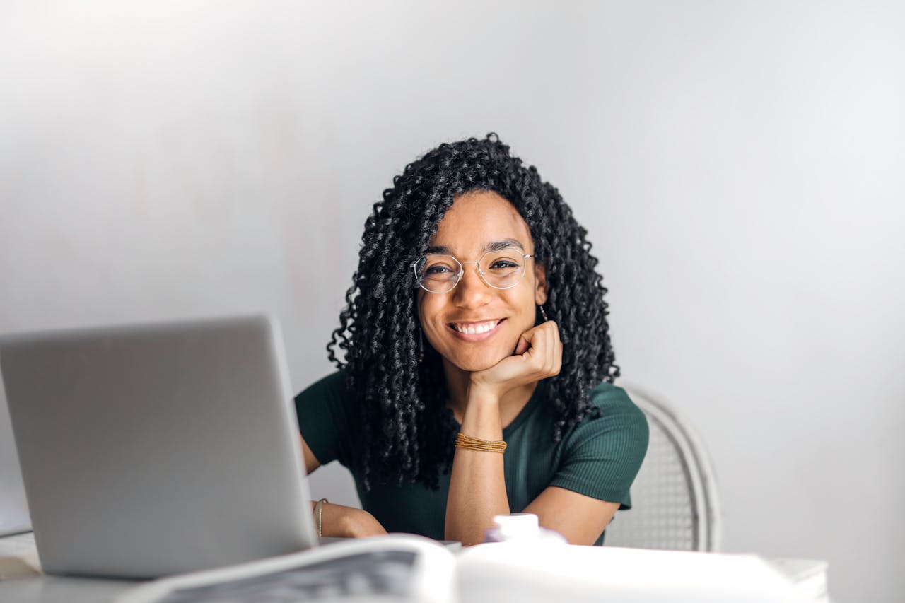 about-us Joyful businesswoman with curly hair smiling at camera while using laptop indoors.